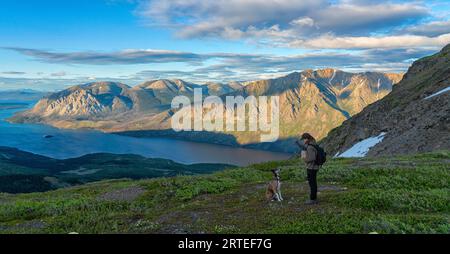 Donne in piedi su una cima di una montagna che dà da mangiare al suo cane e che si godono l'incredibile paesaggio dello Yukon lungo il Sam McGee Trail vicino a Carcross al tramonto Foto Stock