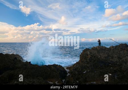 Le onde si infrangono contro la costa vicino al faro Folly mentre un pescatore si trova sulla costa rocciosa; Port Antonio, Giamaica Foto Stock