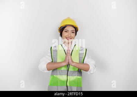 Simpatica lavoratrice asiatica nella fabbrica industriale, saluto tradizionale gestante, indossa un casco di sicurezza giallo, giubbotto verde e uniforme, isolata Foto Stock