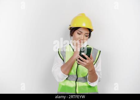 Una giovane lavoratrice premurosa che indossa elmetto di sicurezza e giubbotto mentre tiene il telefono e la mano sul mento, isolata da uno sfondo bianco. Manodopera Foto Stock