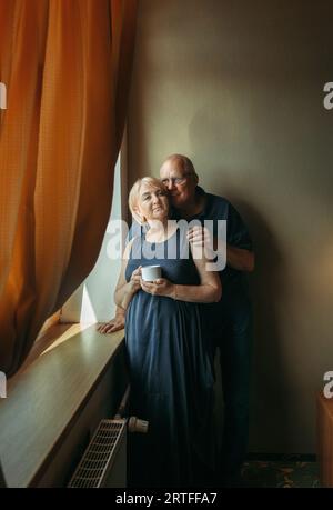 Ritratto di una coppia sposata anziana in piedi alla finestra. L'uomo abbraccia la donna per le spalle. Piano verticale Foto Stock