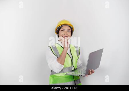 Una giovane lavoratrice premurosa che indossa elmetto di sicurezza e giubbotto mentre tiene il portatile e guarda da parte lo spazio per le copie, isolata da uno schienale bianco Foto Stock