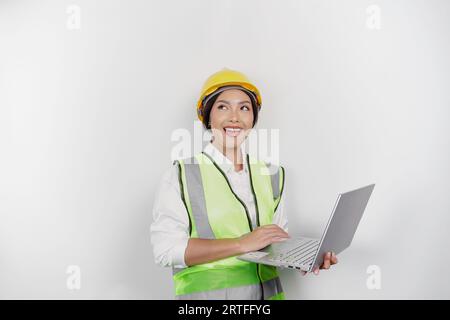 Una giovane lavoratrice premurosa che indossa elmetto di sicurezza e giubbotto mentre tiene il portatile e guarda da parte lo spazio per le copie, isolata da uno schienale bianco Foto Stock