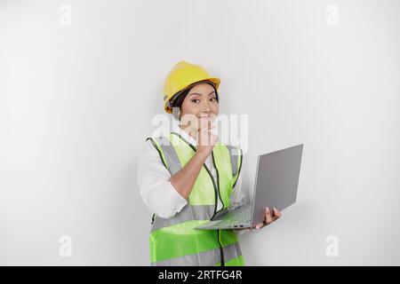 Una giovane lavoratrice premurosa che indossa elmetto di sicurezza e giubbotto mentre tiene il laptop e la mano sul mento, isolata da uno sfondo bianco. Labo Foto Stock