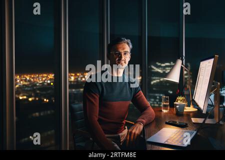 Un uomo d'affari caucasico concentrato che lavora a tarda notte in un ufficio. Si siede alla sua scrivania e guarda la fotocamera mentre lavora diligentemente su un notebook. Foto Stock