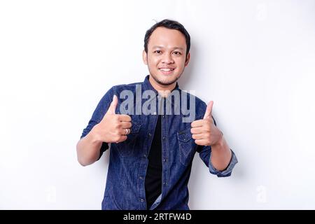 L'eccitato uomo asiatico che indossa una camicia blu dà il pollice su mano gesto di approvazione, isolato da sfondo bianco Foto Stock