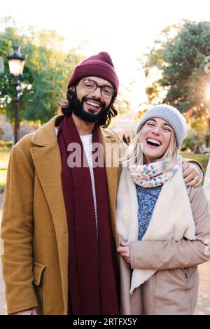 Ritratto verticale di una coppia giovane adulta che sorride e sorride guardando la macchina fotografica, indossando sciarpa, cappelli, cappotti e abiti invernali o autunnali che si legano e si trovano in un parco cittadino all'esterno. Foto di alta qualità Foto Stock