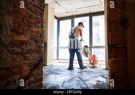 Man che rifinisce la superficie in calcestruzzo con smerigliatrice per massetti in camera con grande finestra. Vista posteriore di un lavoratore maschile che utilizza una saldatrice a conca mentre si lavora sul pavimento dell'appartamento in fase di ristrutturazione. Foto Stock