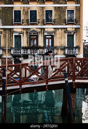 Vecchia signora di colore nero che cammina attraverso il ponte di legno sul canale portando borse per lo shopping davanti alla vecchia casa veneziana Venezia, Italia Foto Stock