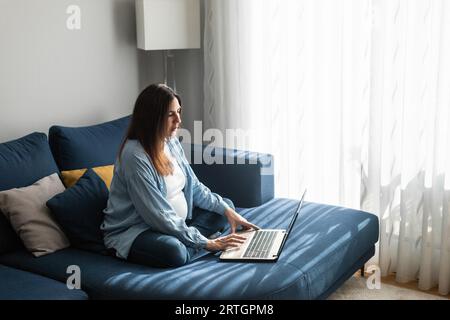 Vista laterale ad alto angolo della donna incinta freelancer seduto divano a casa e navigazione sul netbook mentre si lavora a distanza progetto Foto Stock