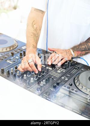 DJ maschio tatuato irriconoscibile in maglietta bianca con controller per regolare l'audio della musica mentre si è in piedi alla console di mixaggio durante la festa in discoteca Foto Stock