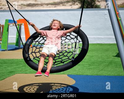 Bambino brillante con capelli ricci dorati in abiti casual d'estate che si diverte nell'area giochi sdraiato sull'altalena del nido con gli occhi chiusi dai raggi che cadono Foto Stock