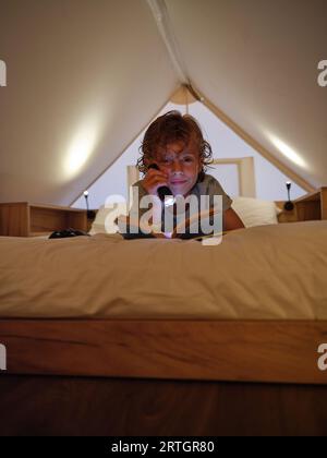 Content boy che illumina il libro e guarda la fotocamera mentre si stende su un comodo letto bianco sulla mansarda a casa Foto Stock