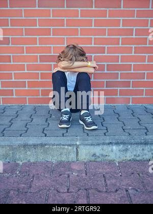 Un bambino preadolescente solitario seduto sul marciapiede vicino a un muro di mattoni sulla moderna strada cittadina di Nazare e abbracciando le ginocchia Foto Stock