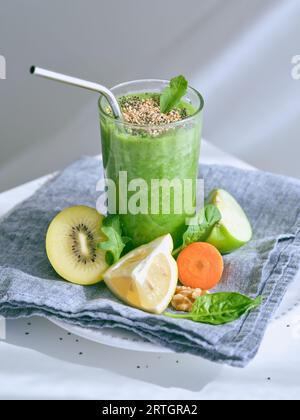 Bicchiere di frullato verde fresco sano con semi di sesamo posto su un asciugamano con abbondanza di frutta tagliata su sfondo bianco Foto Stock