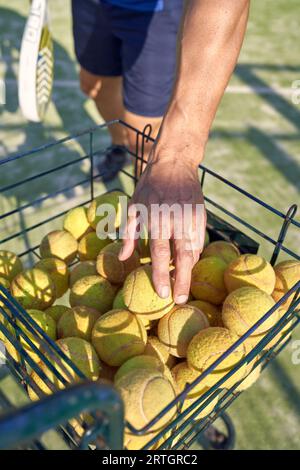 Dall'alto di un atleta maschile irriconoscibile con racchetta da tennis in abbigliamento sportivo che si prepara per l'allenamento in campo e prende la palla gialla dal cestino di metallo Foto Stock