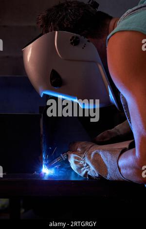 Vista laterale del saldatore maschio irriconoscibile nella struttura metallica di saldatura a casco protettivo durante il lavoro in officina professionale con luce soffusa Foto Stock