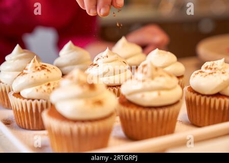 Il coltivatore cosparge la polvere di cannella su gustosi cupcake ricoperti di crema di burro e posti su un tavolo di marmo in cucina a casa Foto Stock