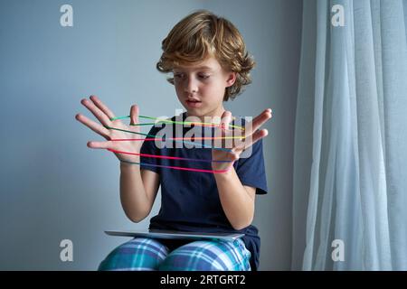 Bambino attento con un abbigliamento casual e un tablet che crea un personaggio con corde colorate mentre gioca a culla di gatti in una stanza luminosa Foto Stock