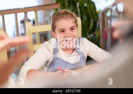 Angolo alto di allegra studentessa con i capelli biondi in abiti casual sorridente mentre ascoltava l'insegnante anonimo seduto al tavolo con no Foto Stock