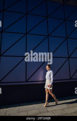 una donna cammina vicino alla parete di vetro di un edificio che cammina per strada Foto Stock