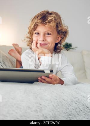 Dal basso di tutto il corpo di un grazioso e affascinante preadolescente con capelli biondi ricci che indossa abiti casual sdraiati sul divano con un tablet e guardano la fotocamera con Foto Stock