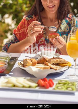 Coltiva una viaggiatrice non riconoscibile che applica marmellata al tavolo con cibi freschi assortiti durante la colazione sulla soleggiata terrazza dell'hotel Foto Stock