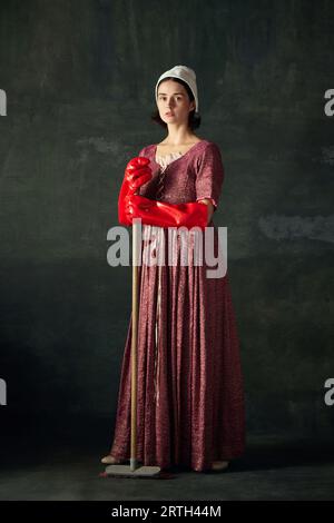 Bella ed elegante donna, cameriera medievale in abito con mop in grandi guanti di gomma rossi su sfondo vintage scuro Foto Stock