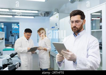 Un giovane scienziato, ingegnere si trova in laboratorio e usa un tablet. Dietro di lui, i colleghi discutono di risultati e ricerche. Foto Stock