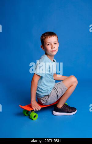 Ragazzo con T-shirt blu mockup seduto sullo skateboard isolato sullo sfondo blu chiaro dello studio Foto Stock