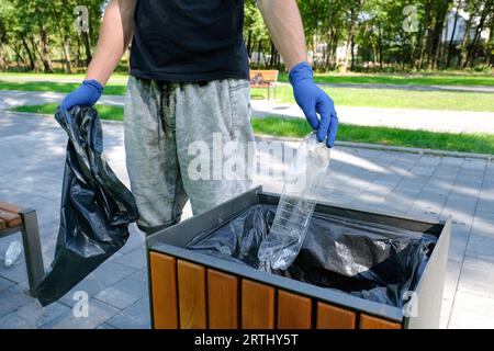 Un volontario maschio raccoglie la spazzatura in un parco pubblico e getta una bottiglia in un cestino. Foto Stock