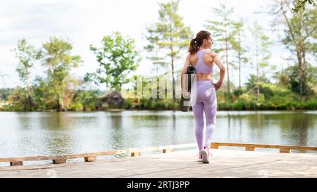 Donna sportiva in piedi su un molo vicino a un lago con in mano un tappetino per esercizi, parte della serie. Foto Stock