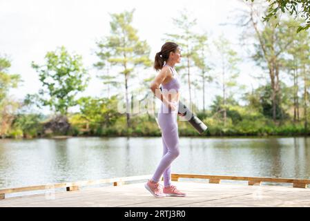 Donna sportiva in piedi su un molo vicino a un lago con in mano un tappetino per esercizi, parte della serie. Foto Stock