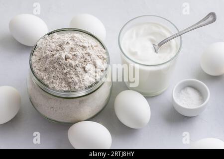 Ingredienti per torte di segale fatte in casa con uovo: Farina di segale, panna acida o yogurt e uova su sfondo grigio chiaro, vista dall'alto, messa a fuoco selettiva Foto Stock