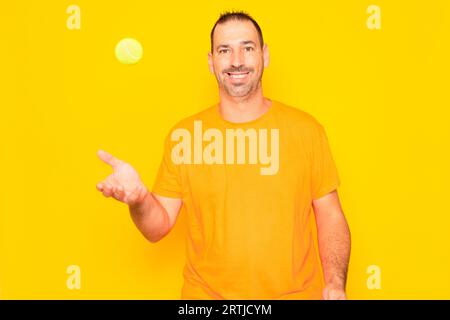 Uomo ispanico con barba negli anni '40 che lancia una palla da tennis nell'aria isolato su sfondo giallo dello studio Foto Stock