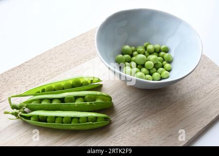 Piselli da giardino (Pisum sativum) in una ciotola di ceramica blu con cialde di pisello, su un tagliere di legno. Isolato su sfondo bianco. Orientamento orizzontale. Foto Stock