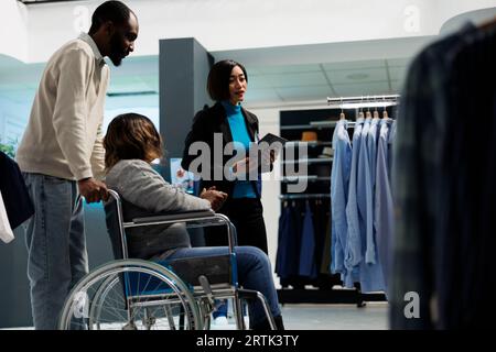 Donna afro-americana con disabilità cronica che riceve consigli da un assistente del negozio di abbigliamento mentre sceglie l'abito. Cliente boutique del centro commerciale in sedia a rotelle per abbigliamento casual Foto Stock