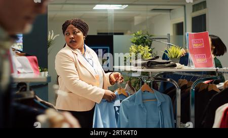 Il lavoratore del punto vendita cerca di calmare le persone pazze che si affrettano a ottenere le migliori offerte sugli articoli di abbigliamento, entusiasma la folla dei clienti in fretta per le promozioni del Black friday. Gli acquirenti afferrano i vestiti. Scatto manuale. Foto Stock