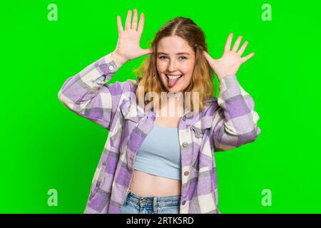 Divertente comica e giocosa giovane donna che fa sciocchezze espressioni facciali e grigia, ingannando, mostrando la lingua, l'espressione idiota. Ragazza isolata su sfondo cromatico verde. Lo stile di vita delle persone Foto Stock