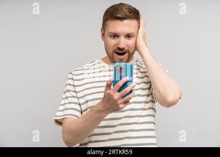 Uomo scioccato che guarda lo schermo dello smartphone stringendo la testa su uno sfondo grigio. Foto Stock