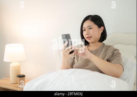 Una bella donna asiatica in abiti comodi che usa lo smartphone, chiacchierando con gli amici mentre si rilassa sul letto della sua camera da letto. Persone AN Foto Stock