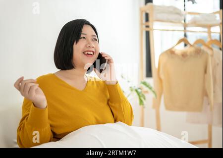 Una bella e felice donna asiatica più grande si diverte a parlare con la sua amica al telefono mentre si siede su un divano e si rilassa nel suo soggiorno. Foto Stock