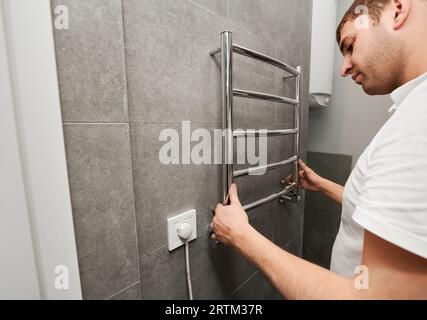 Un lavoratore maschio che appese un portasciugamani elettrico riscaldato alla parete del bagno. Uomo che installa un termoasciugamani in acciaio inox nell'appartamento in fase di ristrutturazione. Concetto di ristrutturazione della casa. Foto Stock