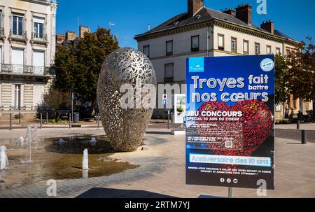 Un cartello che segna il decimo anniversario della scultura conosciuta come il 'cuore di Troyes' accanto a una fontana d'acqua e alle tradizionali case cittadine di Troyes, Aub Foto Stock