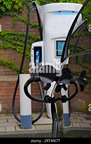 Auto elettrica collegata con cavo e spina alla stazione di ricarica EV per ricaricare la batteria del veicolo, sfondo di una parete con edera hedra per il concetto ecologico Foto Stock