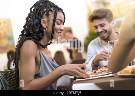 Una donna afro-americana con acconciature afro condivide un momento di gioia affettando la pizza con gli amici caucasici nel cortile di una pizzeria italiana. Caldo, convivia Foto Stock