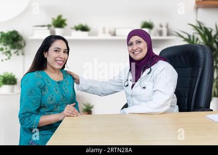 La giovane donna indiana si consulta sul suo problema con una ginecologa musulmana che indossa l'hijab in una clinica moderna. Guardando la fotocamera. Foto Stock