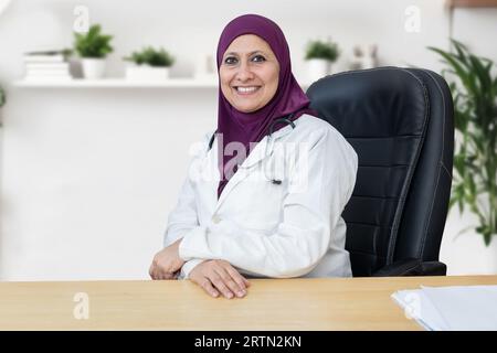 Medico femminile musulmana che indossa hijab che lavora in moderno studio clinico, Ritratto di medico donna araba in cappotto medico bianco con stetoscopio seduto su Foto Stock