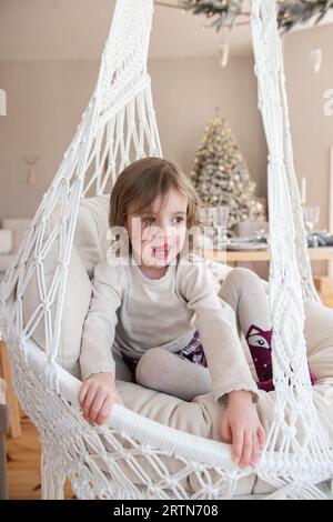 Ritratto ravvicinato di una simpatica bambina su un'altalena di vimini macrame vicino all'albero di Natale a casa. Una bambina allegra sta giocando, la sta tirando fuori Foto Stock