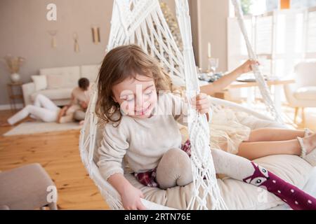 Due bambine cavalcano su un altalena di macrame a casa vicino all'albero di Natale. Le sorelle si divertono, ingannano in vacanza. Ragazze divertenti che hanno un hap Foto Stock